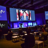 Justin Trudeau apparaît en gros plan sur un écran, devant une assemblée de participants à un forum.