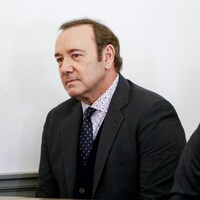 Un homme portant un costume gris et une cravate regarde devant lui. 