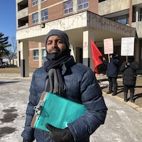 Un homme à l'extérieur devant un immeuble. Il tient dans ses mains des documents. Derrière lui, deux manifestants avec des pancartes.