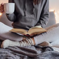 Une femme lit un livre au lit avec une boisson chaude dans une main.