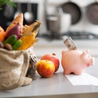 Une tirelire en forme de cochon contient un billet de banque. À côté, un sac en tissu contenant des fruits et des légumes.