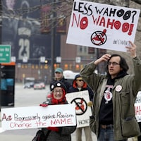 Des gens manifestent en tenant à bout de bras des pancartes sur lesquelles il est inscrit : « Less Wahoo? No Wahoo » et « Act Progressively against Racism. Change the Name ».