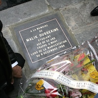 Une personne est agenouillée devant une plaque commémorative sur laquelle a été déposé un bouquet de fleurs. 