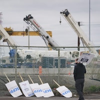 Un homme tient une pancarte près du port de Montréal; d'autres pancartes sont appuyées sur une clôture.