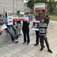 Une dizaine de personnes sont attroupées devant l'Assemblée nationale de Québec et brandissent des pancartes aux couleurs du drapeau iranien, c'est-à-dire vert, blanc et rouge.