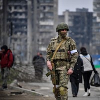 Un soldat russe au milieu d'une rue. En arrière-plan, des gens marchent dans une rue. Des immeubles de logements carbonisés sont visibles. 