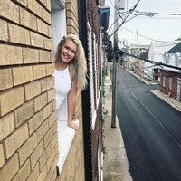 Marylene Levesque a le haut du corps sorti d'une fenêtre, et la vue derrière elle est une rue de la basse-ville de Québec. Elle a les cheveux blonds et est souriante.