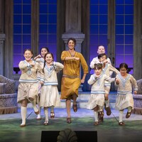 Une femme adulte et sept enfants dansent et chantent dans une scène de la pièce musicale « The Sound of Music ».
