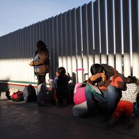 Des enfants et des adultes sont assis au pied d'une clôture avec des valises et des sacs. Un homme se cache le visage de la main.