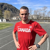Mohamed Aagab debout sur une piste de course extérieure, les mains sur les hanches. Il porte un chandail rouge avec le mot Canada en lettres blanches. On voit la montagne à Campbellton derrière lui.
