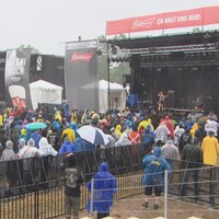 Une centaine de personnes vêtues de manteaux de pluie écoutent un concert rock extérieur, sous la pluie. 