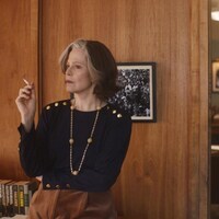 Une femme fume une cigarette, assise sur un meuble dans un bureau. En arrière-plan, on aperçoit une autre femme qui parle au téléphone, assise derrière un bureau.