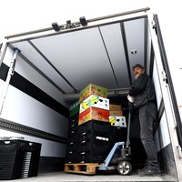 Un homme debout dans un camion tient un diable sur lequel sont empilées des caisses de nourriture.