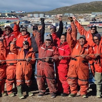 Un groupe de chasseurs vêtus d'une combinaison rouge tiennent des harpon dans leurs mains, devant une vue d'Iqaluit, vendredi 5 août 2022.