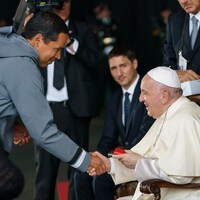 Un homme debout serre la main du pape, assis. Plusieurs personnes autour regardent.