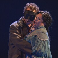 Deux chanteurs d'opéra chantent, dans les bras l'un de l'autre. L'homme a un masque sur les yeux et la femme a les cheveux tressés.