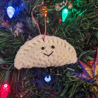 Dans un sapin de Noël tout illuminé, gros plan sur un ornement en laine représentant un pierogi avec deux yeux et une bouche qui sourit.