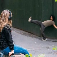 Une femme assise avec des écouteurs regarde un homme effectuer un mouvement de danse dans une ruelle.