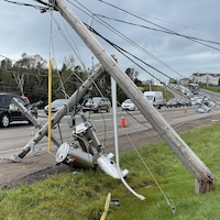Des poteaux électriques brisés et des lignes à haute tension abattues en bordure d'une route sur laquelle des automobiles circulent.