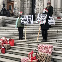 Deux personnes coiffées d'un bonnet de Noël portant des pancartes devant des cadeaux emballés posés sur les marches de l'Assemblée législative.
