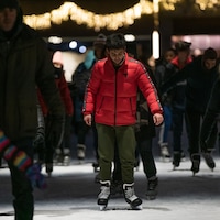 Un jeune homme qui porte un manteau rouge patine parmi d'autres résidents sur une patinoire extérieure. 
