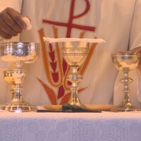 Plan sur les mains du prêtre qui tient, d'une main, des hosties et de l'autre, un verre. 