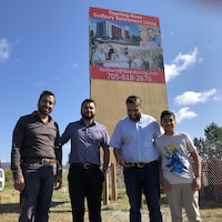 4 hommes posent devant une affiche annonçant une prochaine construction. 