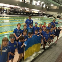 Des enfants devant une piscine olympique brandissent un drapeau bleu et jaune avec leurs entraîneurs.