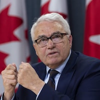 Raymond Théberge, assis devant des drapeaux canadiens, répond à une question, les poings fermés.