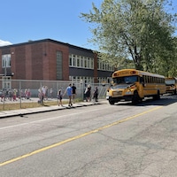 Des autobus scolaires sont stationnés devant une école. Des enfants sont dans la cour.