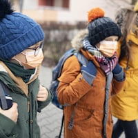 Trois jeunes d'âge différents marchent vers l'école avec des sacs à dos. Ils portent chacun un masque non médical et sont habillés pour le temps froid avec un manteau et une tuque.