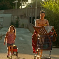 Image du film. Dans une ruelle, trois adultes poussent chacun un panier d'épicerie rempli de chaises, tissus et autres objets, alors qu'une petite fille tire un chariot.