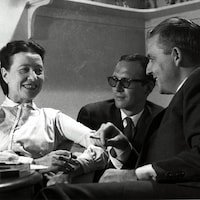 Simone de Beauvoir et Wilfrid Lemoine fumant une cigarette à une petite table. Claude Sylvestre est également assis avec eux, un peu en arrière-plan.