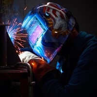 La main gantée du soudeur est à l'oeuvre sur une pièce métallique, les flammèches illuminent le masque flamboyant de l'ouvrier. 