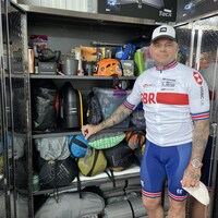 Une homme vêtu d'un ensemble de vélo pose devant une armoire remplis d'équipement d'alpinisme.