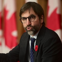 Steven Guilbeault parle devant un micro. Derrière lui sont installés plusieurs drapeaux canadiens.