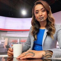 Une femme pose sur un plateau télé avec une tasse de café à la main. On voit un tatouage sur son menton et ses lèvres, ainsi que sur son bras et sa main. 