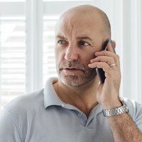 Un homme debout près d'une fenêtre parle au téléphone l'air inquiet.