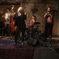 Une femme blonde chante entourée de quatre musiciens de jazz et d'une joueuse de batterie.