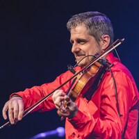 Un musicien sur scène photographié de profil, vêtu d'une chemise rouge et jouant du violon.