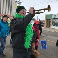 Une personne porte une écharpe à plumes vertes et souffle à la trompette.