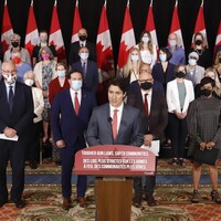 Justin Trudeau, entouré de personnes qui portent des masques, devant des drapeaux et devant un lutrin.