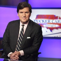 Tucker Carlson pose pour des photos dans un studio de la chaîne Fox News, devant un écran où on voit le titre de l'émission qui porte son nom.