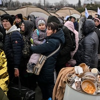 Des réfugiés, en habits et manteaux d'hiver, attendent en file. Autour d'eux, des tentes et des déchets.