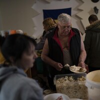 Un homme dans la soixantaine se sert un repas au milieu d'autres réfugiés dans une pièce remplie de sacs et d'effets personnels.