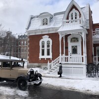 La photo montre une maison victorienne, devant laquelle est garée une vieille voiture beige.