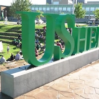Derrière de logo de l'université, des étudiants suivent un cours en plein air.