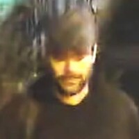 Une capture d'écran de l'homme lors des évènements, il portait une barbe, une casquette, un manteau noir, un pantalon noir, un sac à dos et des bottillons beiges.