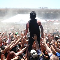 Un chanteur est debout, au dessus d'une foule de spectateurs qui tentent de le toucher.