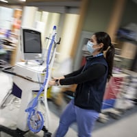 Une femme en uniforme bleu d'hôpital pousse un respirateur artificiel sur roulettes dans une salle.